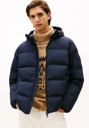 Giovane uomo con un puffer blu navy sopra un maglione beige a colletto alto con un motivo, con le mani in tasca su uno sfondo bianco.