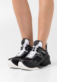 Černo-bílé sportovní tenisky s vrchním dílem z prodyšné síťoviny, masivní podrážkou, kontrastními texturami a značkovými akcenty po straně.