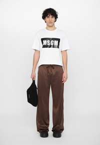 White t-shirt with black "MSG" logo, brown satin pants with drawstring, black pouch, and black clogs, standing on a light background.