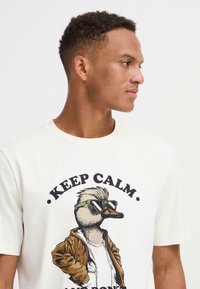 Cremefarvet T-shirt med en tegneserieand, der har grønne solbriller og en brun jakke, med teksten "BEVAR ROEN" over illustration.