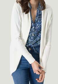 Cardigan blanc à ouverture devant avec texture côtelée, porté sur un chemisier bleu à motif floral et un jean bleu foncé, présentant une coupe décontractée.