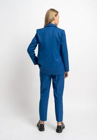 Blauer maßgeschneiderter Anzug mit einem strukturierten Blazer und schmal geschnittenen Hosen, die eine glatte Textur und ein minimales Design aufweisen. Schwarze Schuhe runden den Look ab.