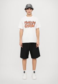 T-shirt alb cu text grafic portocaliu, asortat cu pantaloni scurți negri. Pantofi sport albi cu accente negre. Șapcă cu un logo circular în gri.