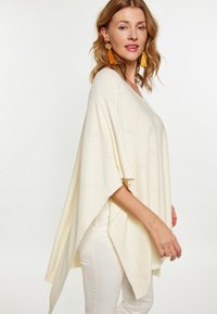Poncho din crem cu model de tricotaj deschis, mâneci largi și design în formă de v. Asortat cu pantaloni albi, care au accente texturate.