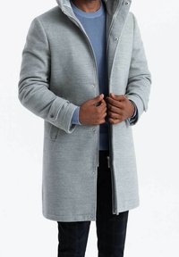 Abrigo acolchado gris con capucha, botones a presión, puños acanalados y bolsillos. Se lleva sobre un suéter azul, mostrando las manos sujetando el cierre.