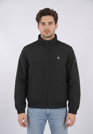 Jeune homme aux cheveux courts et bouclés portant une veste noire zippée avec un logo cK sur la poitrine et un jean bleu clair, debout devant un fond uni.