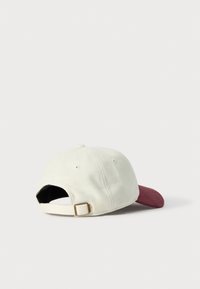Hvid justerbar baseballcap med bordeauxfarvet buet skygge, set fra bag på en simpel lys baggrund.