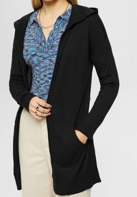 Svart långärmad cardigan med huva, med en lös passform, sidofickor och en mjuk textur, som bärs över en blå mönstrad topp.