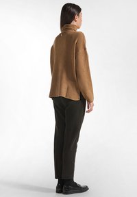Brauner übergroßer Pullover mit hohem Kragen und strukturiertem Finish, kombiniert mit gestreiften dunklen Hosen und glänzenden schwarzen Schuhen.