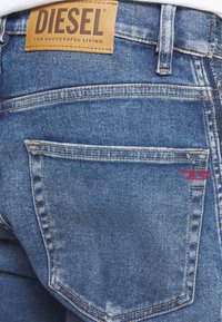 Modré džínové kalhoty z denimu s koženou značkou s nápisem "DIESEL". Na zadním kapse mají červené prošívání a klasický design s pěti kapsami.