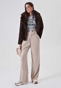 Donna che indossa un giubbotto di pelliccia marrone scuro, una maglietta grigia con grafiche, pantaloni beige a gamba larga, sneaker bianche, mentre tiene una borsa beige, in piedi su uno sfondo neutro.