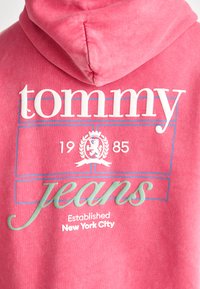 Rozā kapuce ar teksturētu virsmu. Iekļauj balto "tommy" logo, zaļos "džinsus" un grafiku ar lauvu, uz kuras ir "1985" un zila malējā apmale.