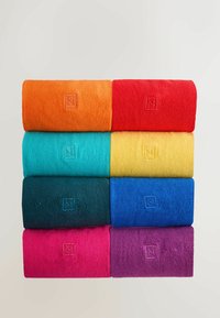 Stos ośmiu zwiniętych ręczników w różnych kolorach: pomarańczowy, czerwony, turkusowy, żółty, ciemnozielony, niebieski, różowy i fioletowy. Każdy ręcznik ma subtelnie wytłoczony logo.