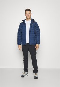 Giacca a vento in blu navy scuro con trapuntatura orizzontale e cappuccio, abbinata a pantaloni neri e scarpe sportive con dettagli grigi e arancioni.