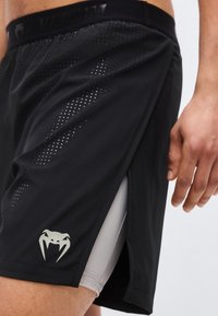 Μαύρα αθλητικά σορτς με διάτρητους πλαϊνούς αεραγωγούς και γκρι κοντά σορτς συμπίεσης από κάτω, εμφανιζόμενα στον μηρό και το χέρι ενός ατόμου.