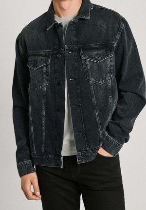 Mann trägt schwarze Jeansjacke mit knöpfbarem Vorderteil und Klappentaschen, kombiniert mit hellgrauem Hemd und schwarzer Hose.