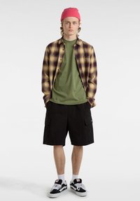 Camisa de cuadros en amarillo, morado y negro, superpuesta a una camiseta verde, combinada con pantalones cortos cargo negros y zapatillas negras con acentos blancos.