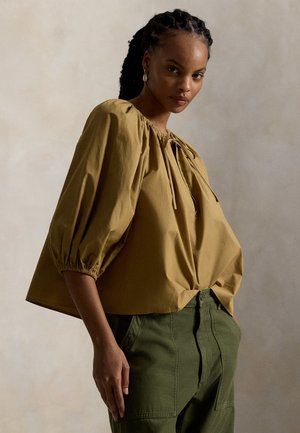 Garstyčių geltonumo palaidinė su pūstomis rankovėmis ir kaklaraiščiu prie kaklo, pasižyminti laisvu siluetu, tekstūruota medžiaga ir surinktomis rankogaliais, derinama su alyvuogių žalios spalvos kelnėmis.