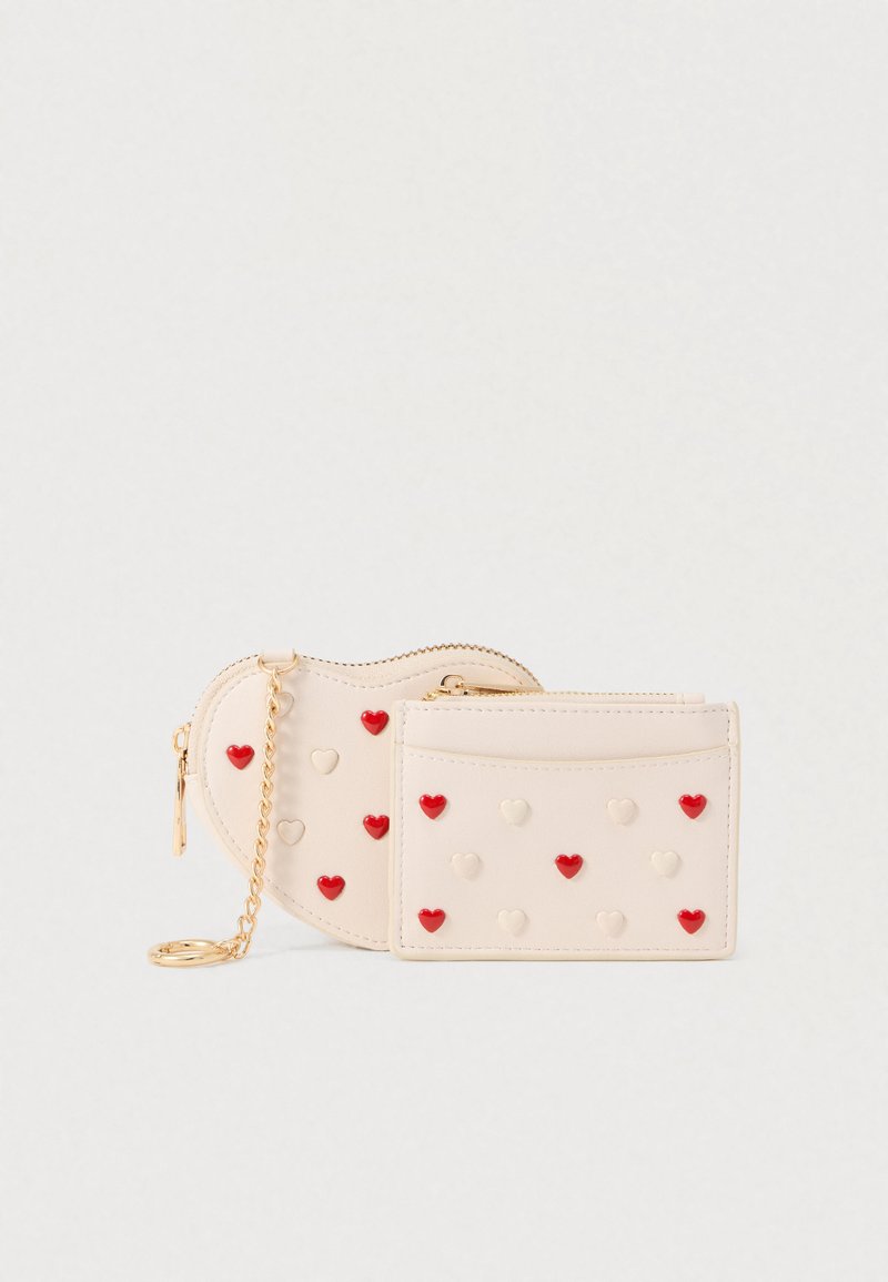 Portefeuille beige compact orné de motifs en forme de cœur rouges et crème. Il possède une fermeture éclair et une chaîne dorée pour un transport facile.