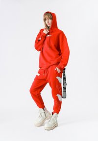 Persona vistiendo una sudadera con capucha roja brillante y pantalones deportivos a juego con parches de alas, botas blancas con cordones, de pie con la mano cerca de la cara.