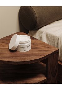 Barattolo di crema per il corpo circolare bianco, parzialmente aperto, su un tavolino in legno scuro. Texture liscia, etichetta minimalista, vicino a un cuscino rivestito di morbido tessuto.