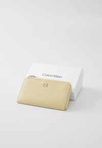 Portefeuille en cuir jaune Calvin Klein avec logo argenté, fermeture éclair et texture lisse, présenté sur une boîte blanche avec impression de logo.