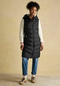 Czarna ocieplana kamizelka o przylegającym kroju, z kapturem i kieszeniami na zamek. Charakteryzuje się gładką teksturą i wzorem w zygzak. W zestawie z niebieskimi dżinsami i sneakersami.