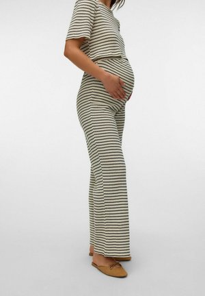 Gravid person iført beige og sort stribet strik top og bukser, holder maven, står med brune flade sko på ensfarvet baggrund.