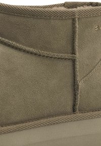 Botín de ante verde oliva con textura suave, detalles de costura y un acento de tela marrón claro en el borde superior. Diseño liviano y de perfil bajo.