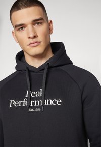 Sudadera con capucha negra de tela texturizada. Presenta texto bordado en blanco "Real Performance" y "Est. 1986", además de acentos de cordón.