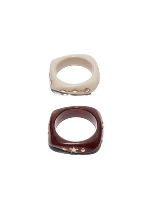 Dve štvorcové prsteňové živice, jeden béžový so zlatými diamantovými tvarmi, druhý tmavohnedý so zlatými hviezdami a okrúhlymi cvočkami, na bielom pozadí.