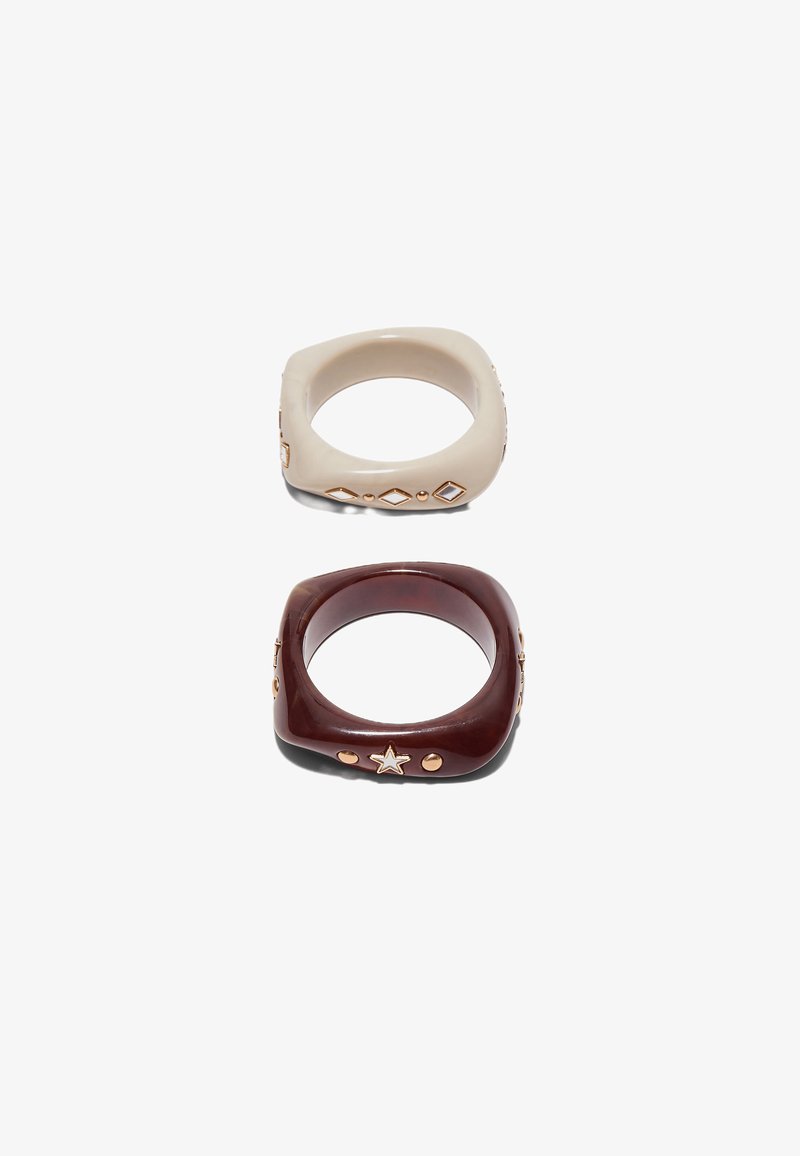 To firkantede ringe i harpiks, den ene beige med gyldne diamantformer, den anden mørkebrun med gyldne stjerne- og runde nitter, på hvid baggrund.