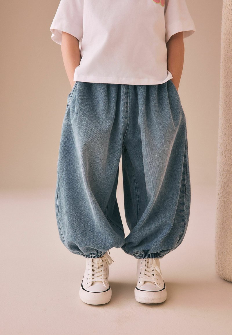 Niño con pantalones vaqueros azules holgados con puños elásticos y zapatillas blancas, de pie con las manos en los bolsillos contra un fondo neutro.