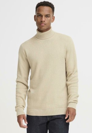Pull en maille beige clair avec un col montant, un motif texturé et des poignets et un ourlet côtelés, associé à un jean bleu foncé.