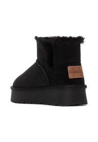 Botas negras hasta el tobillo de ante con un suave collar de piel sintética. Presentan un parche de cuero marrón con el nombre de la marca y una suela gruesa negra.