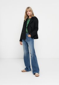 Svart dobbelbreasted peacoat, grønn mønstret genser, lyseblå flagrende jeans, og brune sko med designdetaljer. Glatte stoff teksturer.