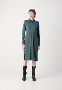 Robe à boutonnière en tissu vert foncé, avec des manches longues, un col et des fentes sur les côtés. Portée avec de grandes bottes de cow-boy noires.