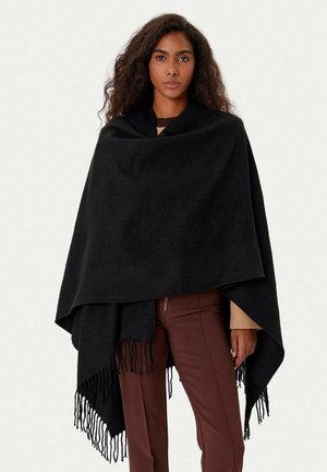 Femme aux cheveux bouclés foncés porte un châle noir à franges par-dessus un top beige et un pantalon marron, debout contre un fond clair uni.