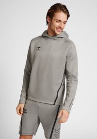 Haut de survêtement en gris avec capuche, manches longues, accents noirs et poche zippée. Associé à un short assorti avec un design rayé et un logo.