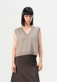 Giovane donna con capelli neri lisci che indossa un top a maglia beige senza maniche con scollatura a V e una gonna marrone scuro, in piedi contro uno sfondo neutro.