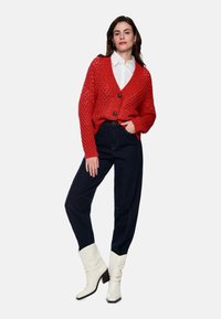 Roter Strickcardigan mit V-Ausschnitt, Knopfleiste und offenem Strickmuster, kombiniert mit dunklen, hoch taillierten Jeans und weißen Stiefeln mit Absatz.