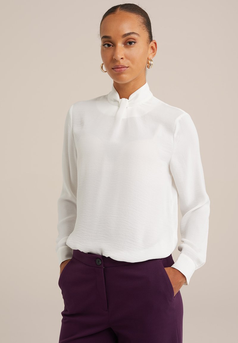 Vit blus med hög krage och långa ärmar, tillverkad av ett lätt tyg, ihop med mörklila skräddarsydda byxor med en knappdetalj.