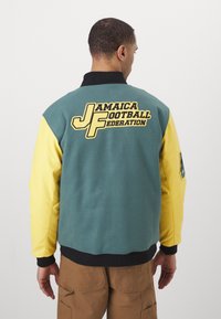Colorful two-tone fleece jacket featuring a teal body and yellow sleeves. Back displays "Jamaica Football Federation" in bold lettering.