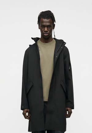 Letitettyä tukkaa oleva mies pukeutuneena mustaan hupulliseen takkiin ja beigen väriseen pyöreäkaulaiseen neulepaitaan, seisoo yksinkertaisen vaaleanharmaan taustan edessä.