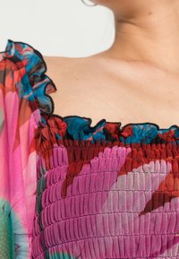 Bunte, durchsichtige, geraffte Bluse mit Rüschensaum, die eine Mischung aus rosa, rot und blauen Mustern auf einem strukturierten Stoff zeigt.