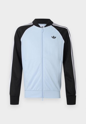 Hellblaues Sportjacke mit schwarzen Ärmeln, weißen Streifen entlang der Ärmel, Reißverschluss vorne und einem kleinen schwarzen Adidas-Logo auf der Brust.