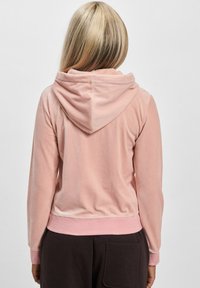 Ružičasti velur dukserica sa mekom teksturom, s kapuljačom na vezanje i rebrastim manžetama, u kombinaciji sa tamnim hlačama. Prikazan zadnji pogled.