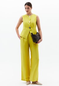 Gilet giallo su misura con tre bottoni, abbinato a pantaloni a gamba larga coordinati. Borsa a tracolla nera intrecciata e scarpe flat decorate.