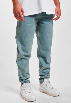 Homme portant un jean en denim bleu clair, des baskets blanches et un t-shirt blanc, debout les mains détendues le long du corps.