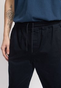 Pantaloni neri con vita elastica e coulisse, caratterizzati da una texture liscia e tasche laterali, indossati con una camicia blu.